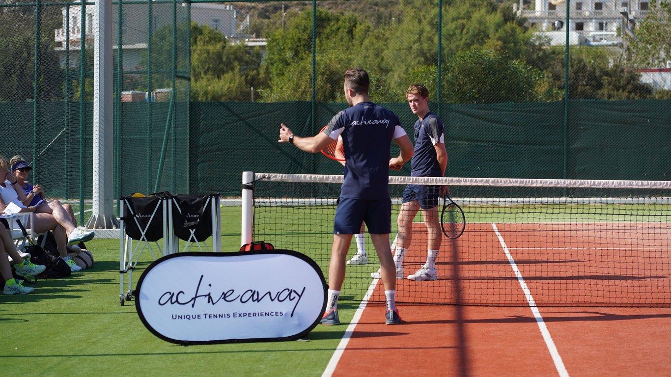 Coach instructs players at the net during an Active Away tennis clinic with the group watching courtside.