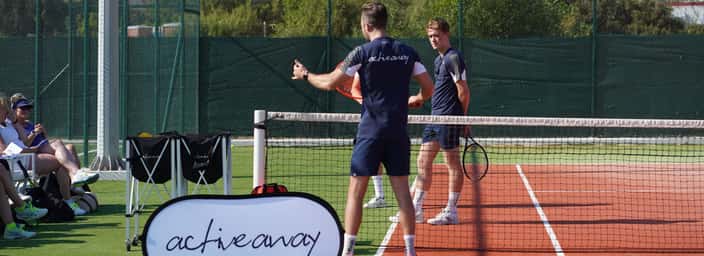 Coach instructs players at the net during an Active Away tennis clinic with the group watching courtside.