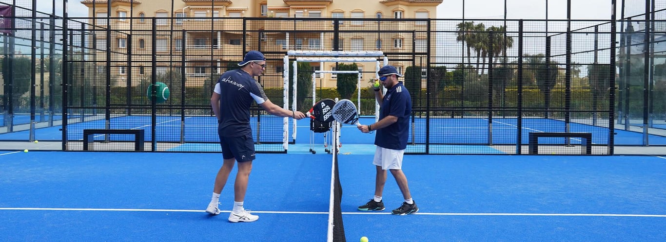 Two padel players tap rackets over the net on a blue court during a holiday coaching session.