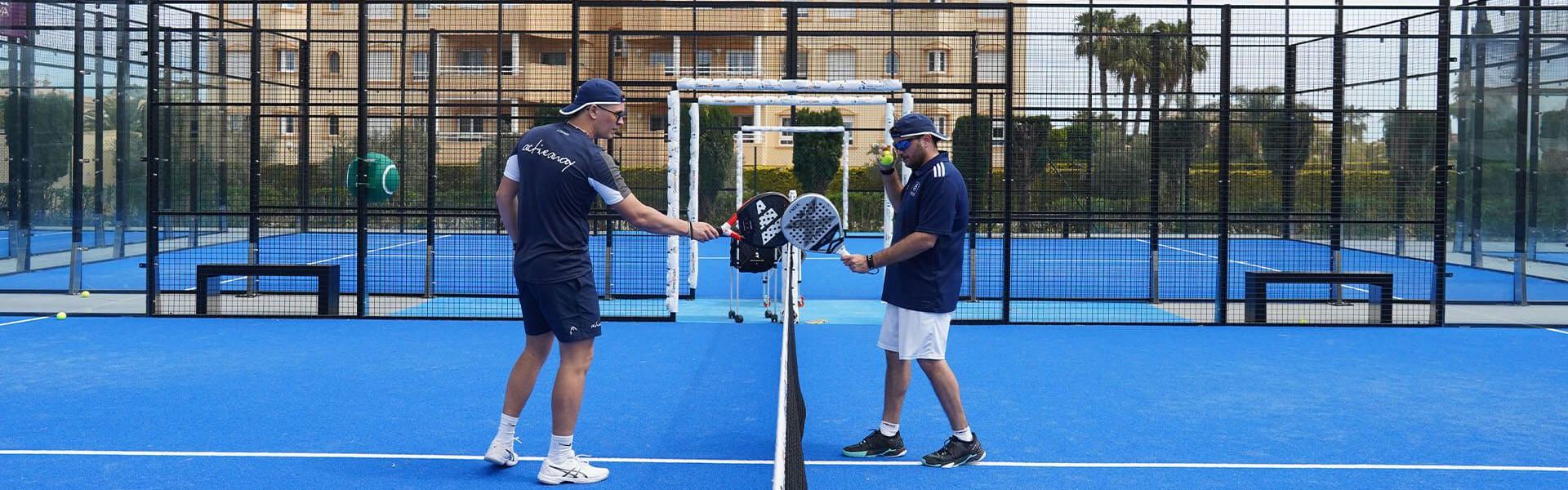 Two padel players tap rackets over the net on a blue court during a holiday coaching session.