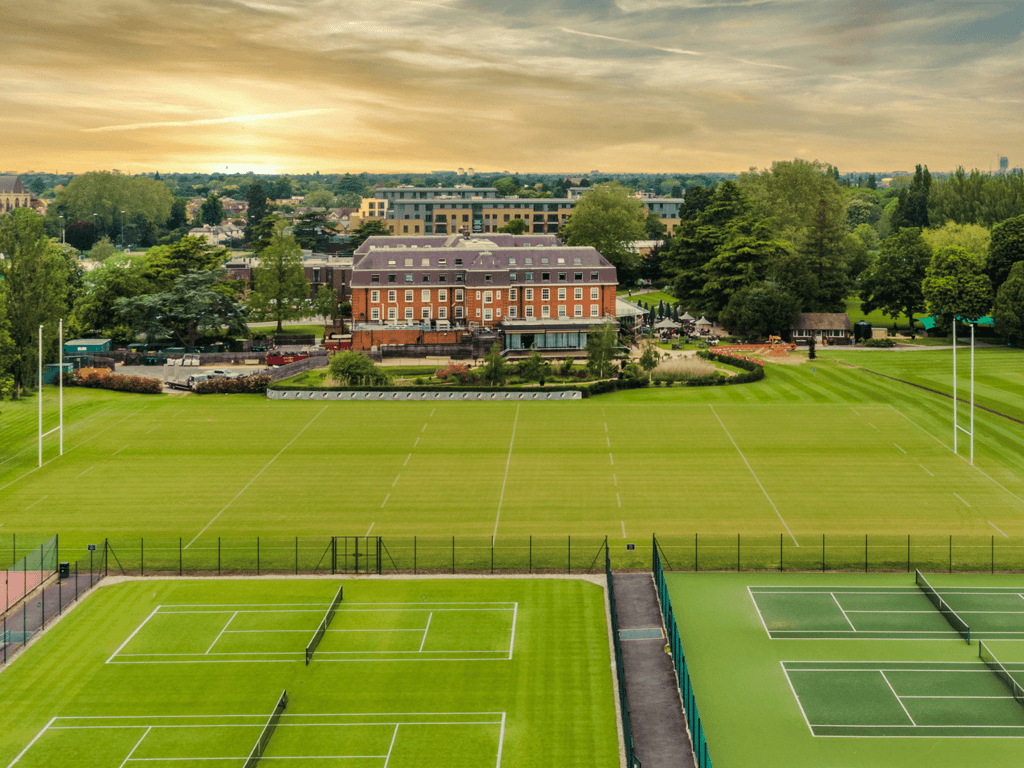 Tennis courts and rugby pitches by a grand clubhouse at sunset, set in lush grounds.