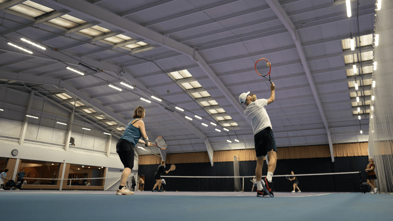 Players practise at an indoor tennis clinic, hitting volleys under bright lights on adjacent courts.