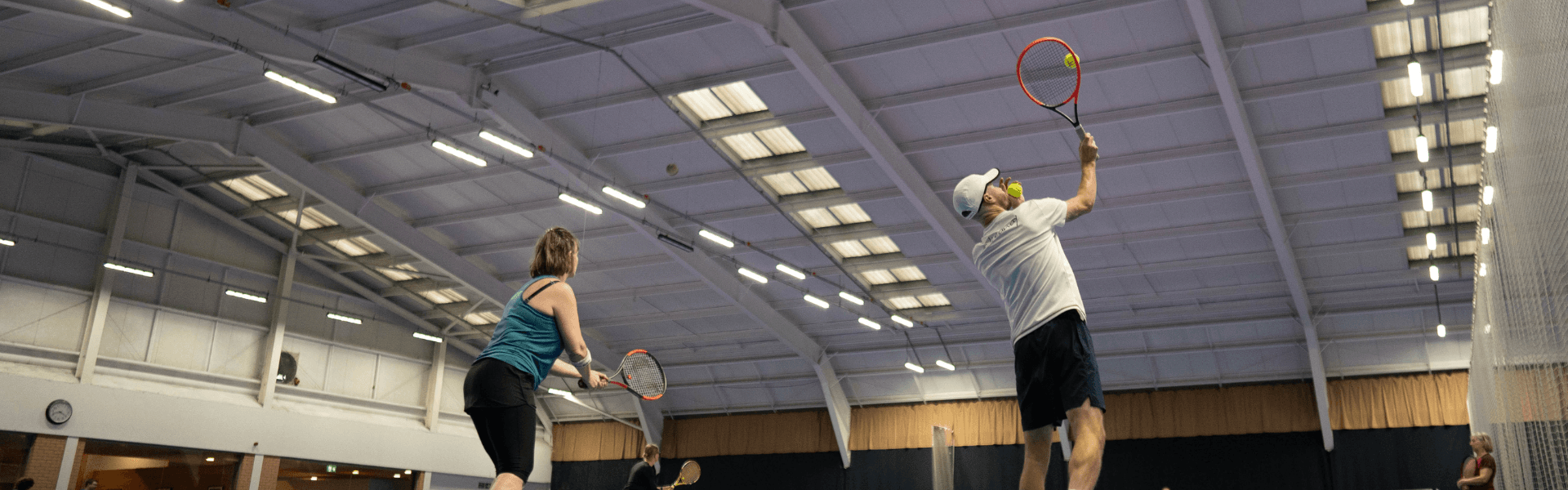 Players practise at an indoor tennis clinic, hitting volleys under bright lights on adjacent courts.