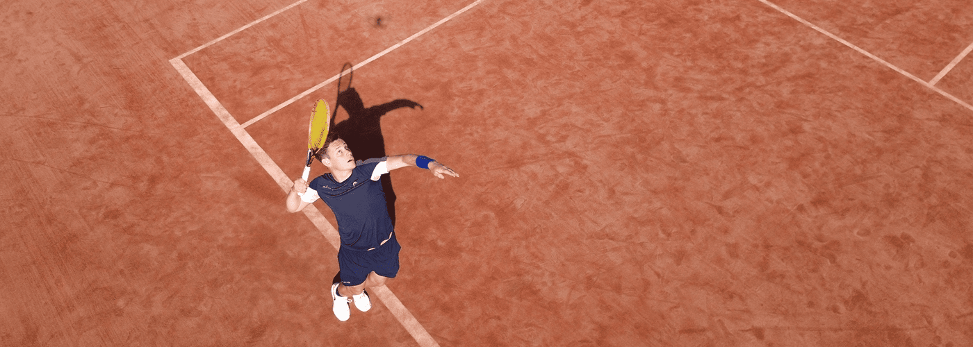 A tennis player leaping to serve on a clay court, seen from above.