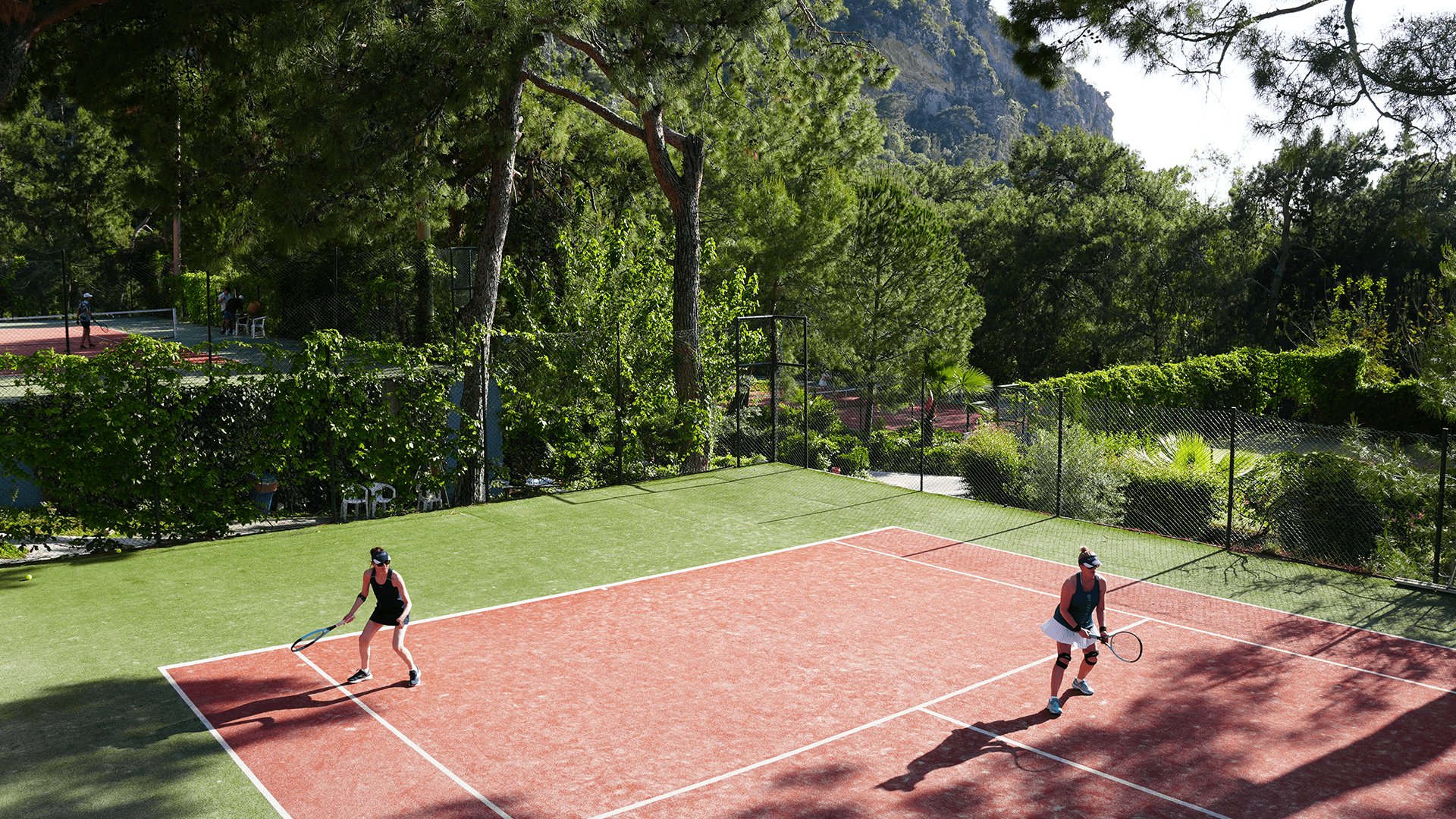 Two players on an outdoor tennis court practising under pine trees at a sunny resort.