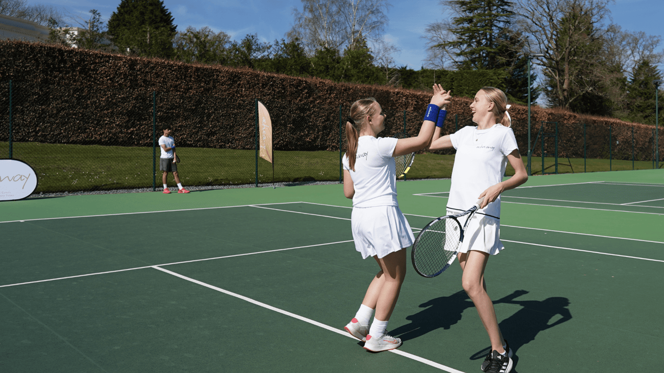Two junior tennis players high-five on an outdoor hard court, racquets in hand, with another child in the background.