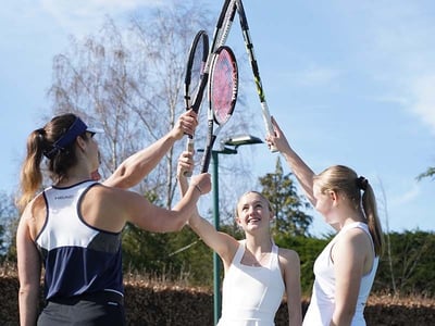 Three junior tennis players raise rackets together in a team huddle on an outdoor court.