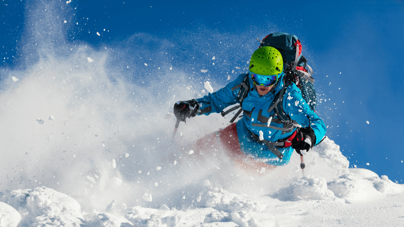 Skier carving through deep powder, snow spraying into the air under a clear blue sky.