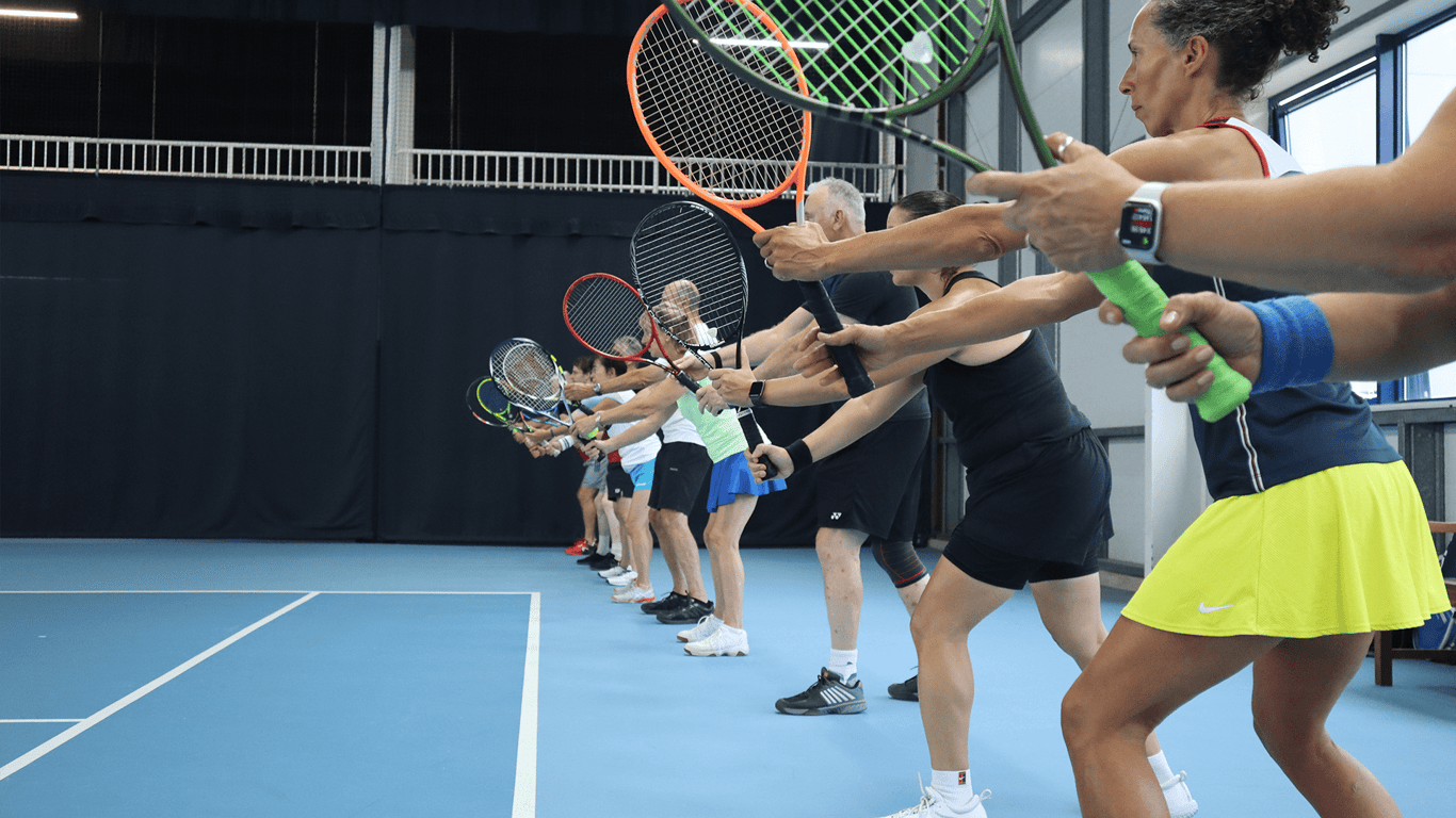 Adults lined up on an indoor court practising forehand swings at a tennis clinic.