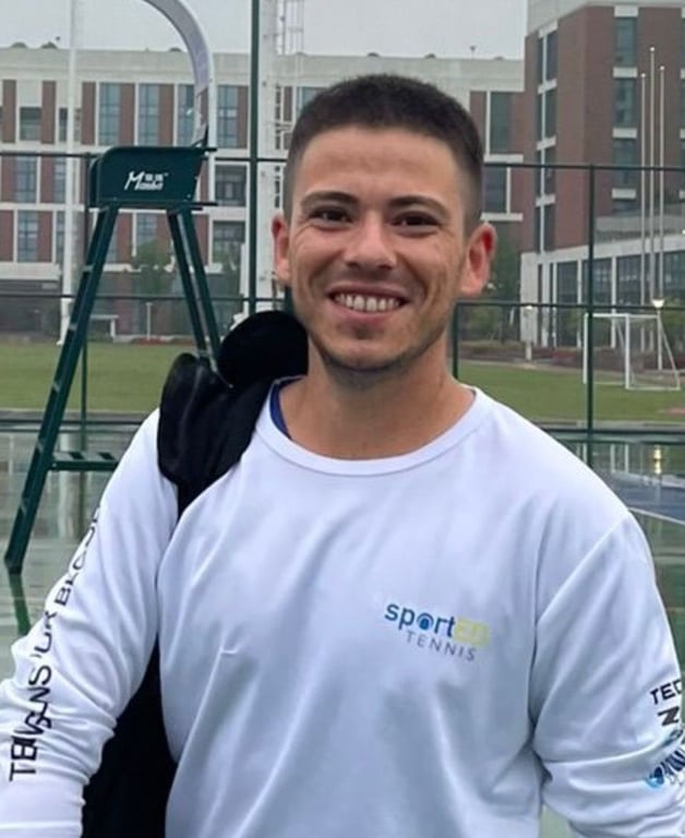 Smiling tennis coach in white top with kit bag on outdoor courts, ready for a session.
