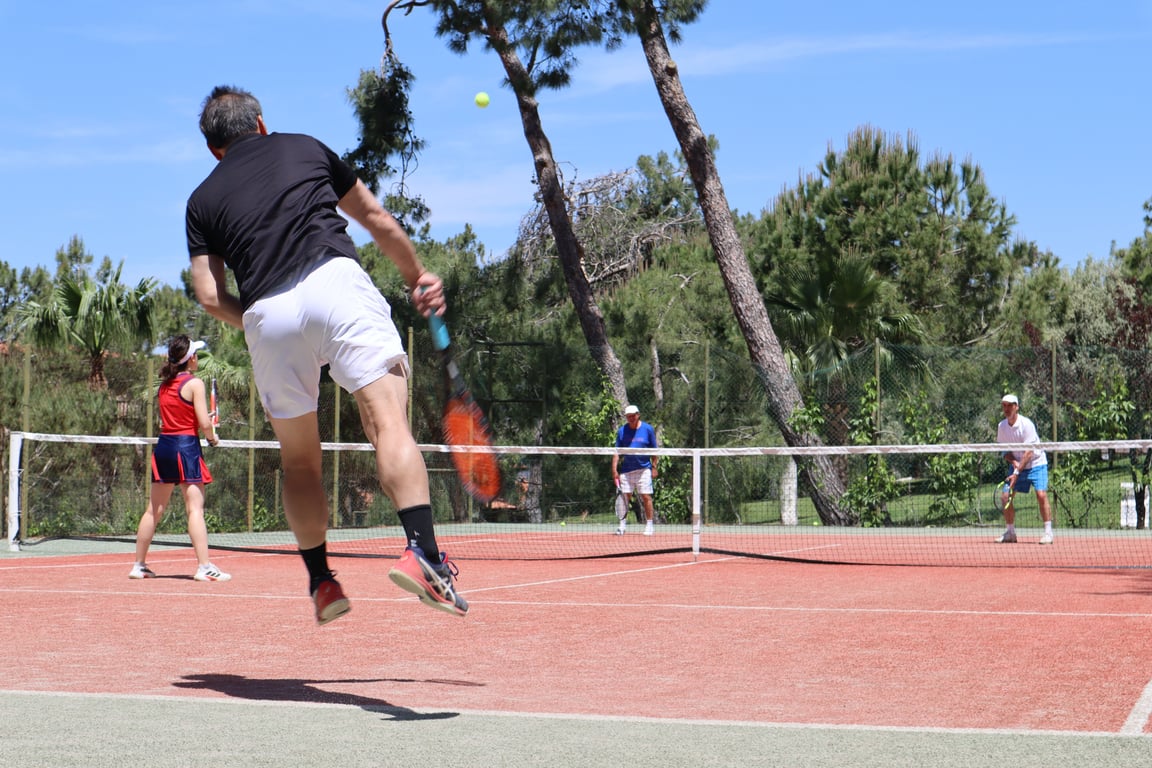 Man jumping to serve in an outdoor doubles tennis match under a sunny sky.