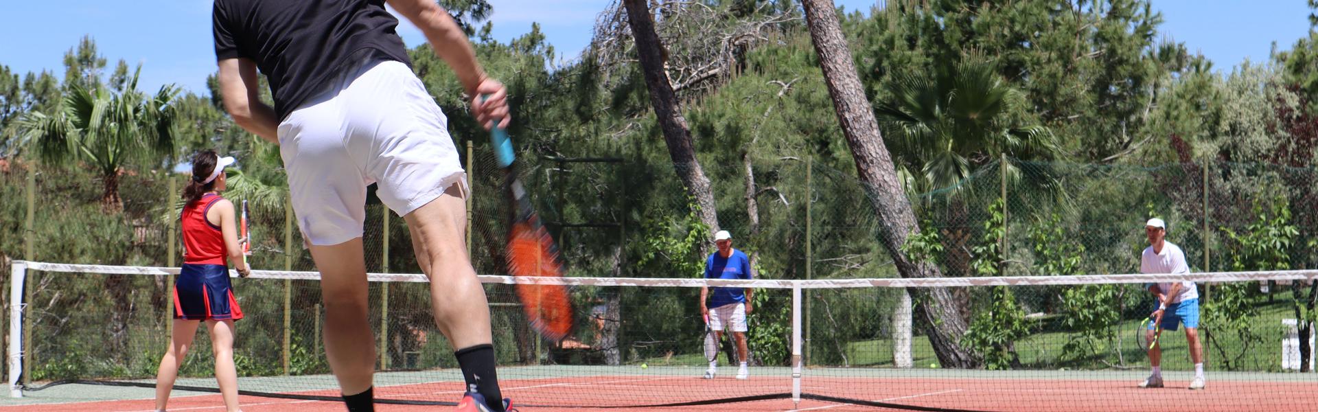 Man jumping to serve in an outdoor doubles tennis match under a sunny sky.