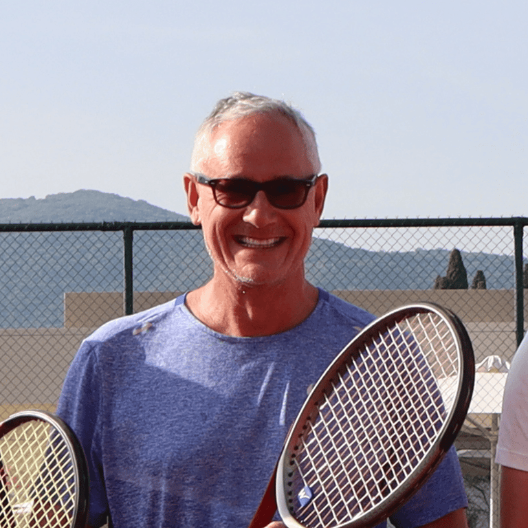 Smiling man in sunglasses holding a tennis racket on an outdoor court.