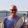 Smiling man in sunglasses holding a tennis racket on an outdoor court.
