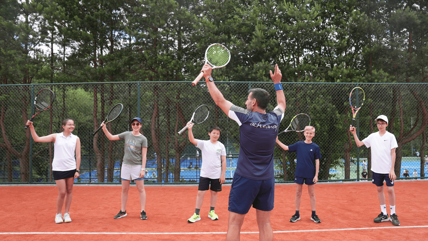 Coach leads juniors raising tennis racquets on a clay court, cheering at a summer camp.