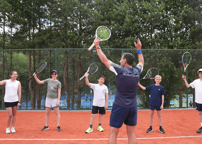 Coach leads juniors raising tennis racquets on a clay court, cheering at a summer camp.