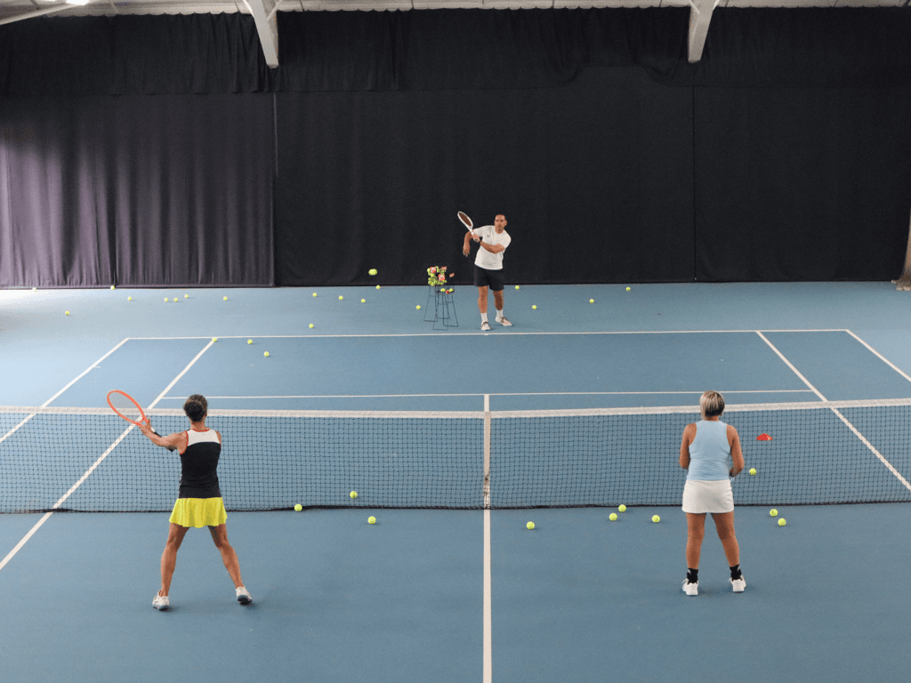 Indoor tennis lesson with coach feeding balls to two players at the net.