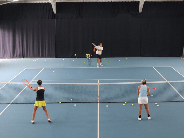 Indoor tennis lesson with coach feeding balls to two players at the net.