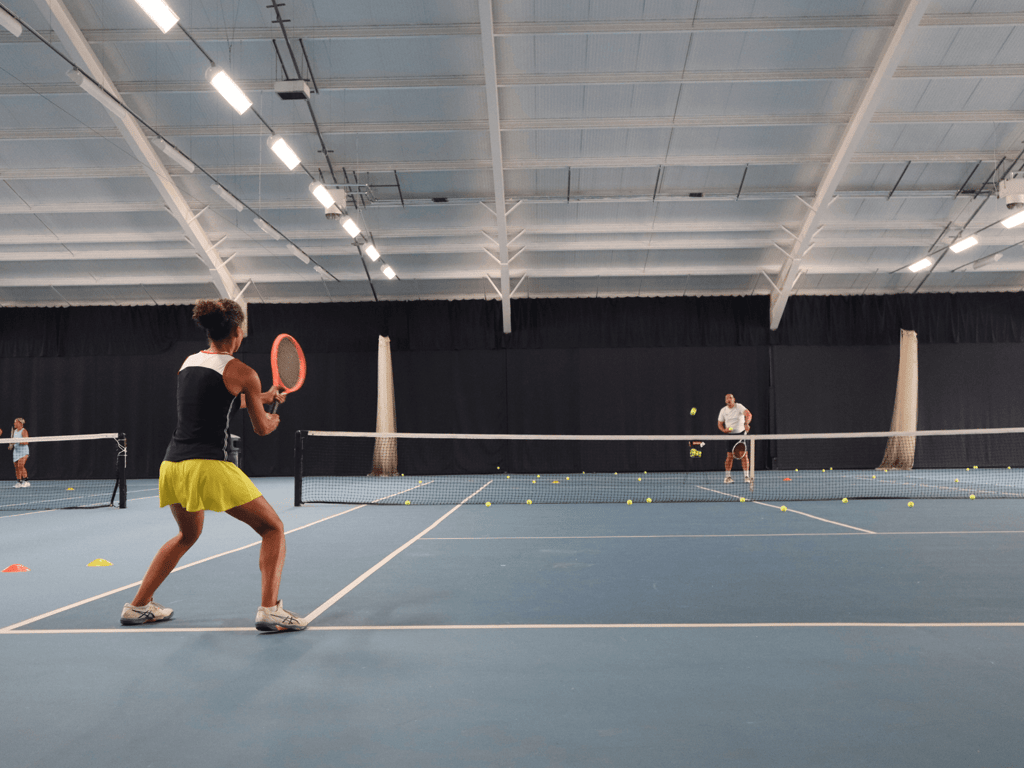 Woman prepares a backhand during indoor tennis training as a coach feeds balls across the net.