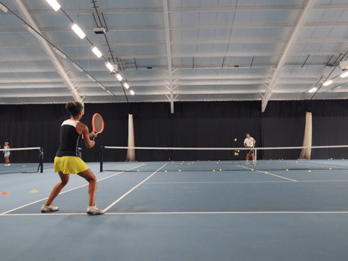 Woman prepares a backhand during indoor tennis training as a coach feeds balls across the net.