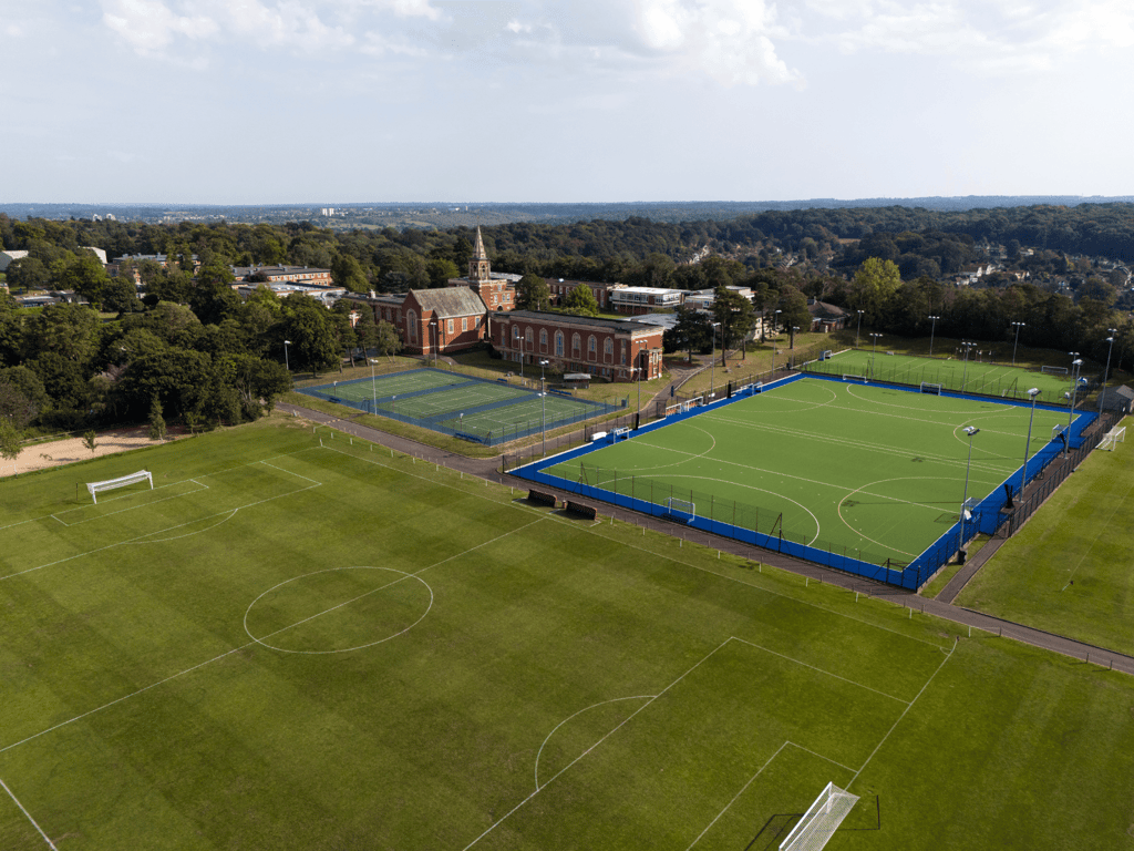 Aerial view of sports complex with blue tennis courts, hockey astro and football pitches by a campus building.