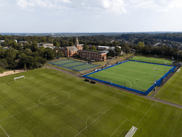 Aerial view of sports complex with blue tennis courts, hockey astro and football pitches by a campus building.
