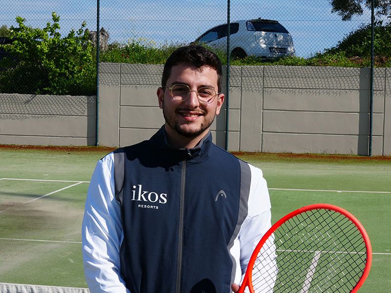 Tennis coach with red racket on outdoor court at Ikos Oceania resort.