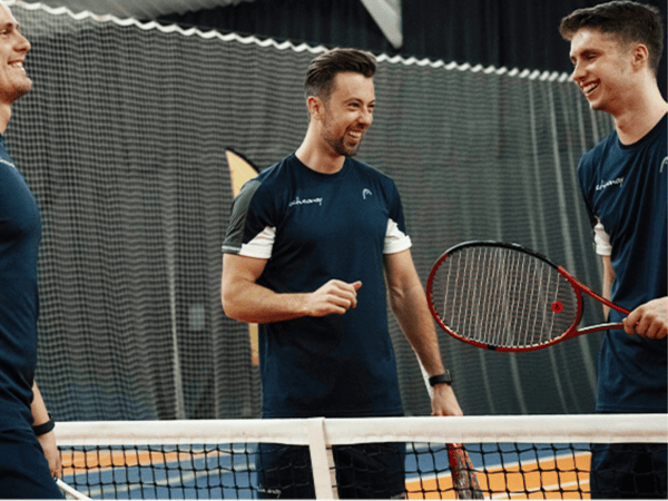 Three men in navy sportswear chatting by a tennis net on an indoor court, one holding a racquet.