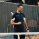 Three men in navy sportswear chatting by a tennis net on an indoor court, one holding a racquet.