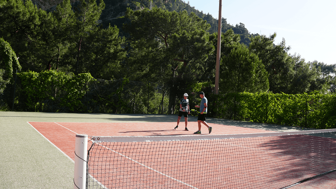 Two players stand on an outdoor tennis court, chatting by the baseline with the net in foreground and trees behind.