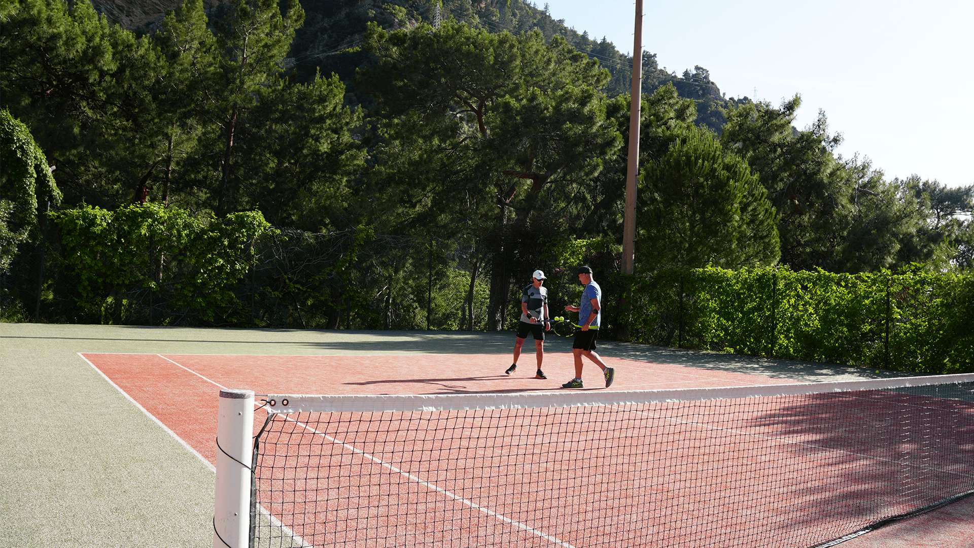 Two players stand on an outdoor tennis court, chatting by the baseline with the net in foreground and trees behind.
