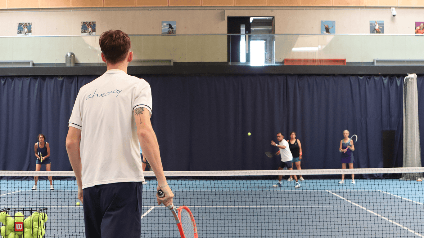 Coach leads indoor tennis drill as group of players practise across the net.