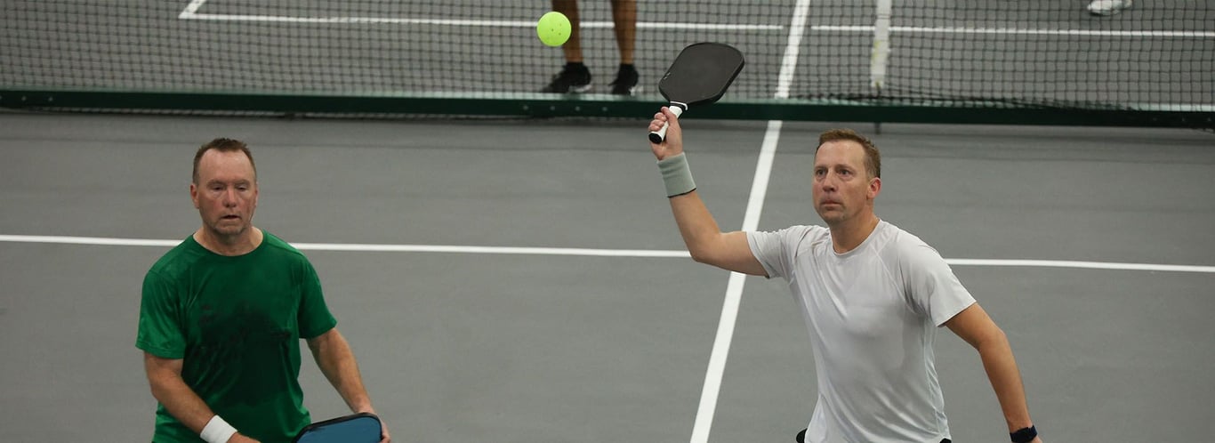 Two men play doubles pickleball indoors; one in white prepares to hit a high ball with a paddle.
