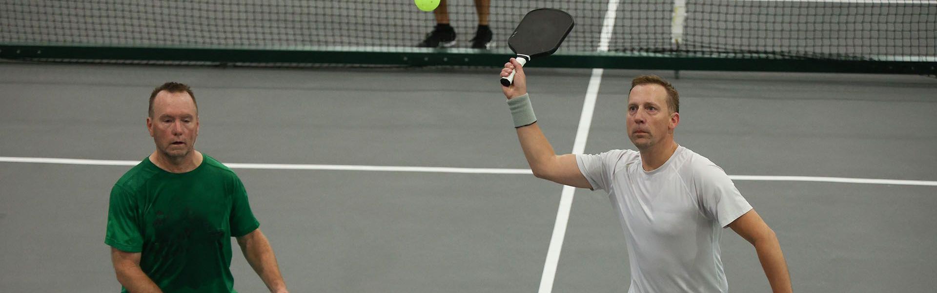 Two men play doubles pickleball indoors; one in white prepares to hit a high ball with a paddle.