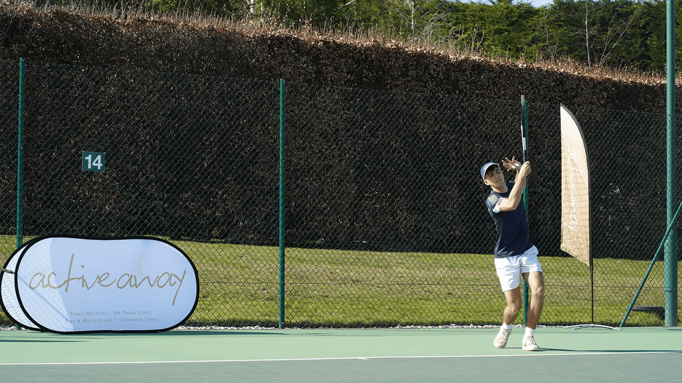 Tennis player serving on an outdoor court with Active Away branding on a sunny day.