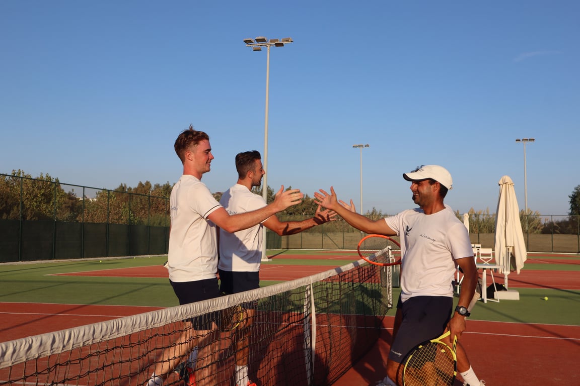 Tennis coaches exchange handshakes at the net after a doubles session on a sunny outdoor court.