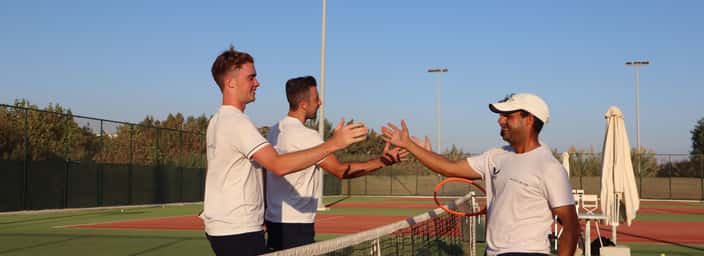Tennis coaches exchange handshakes at the net after a doubles session on a sunny outdoor court.