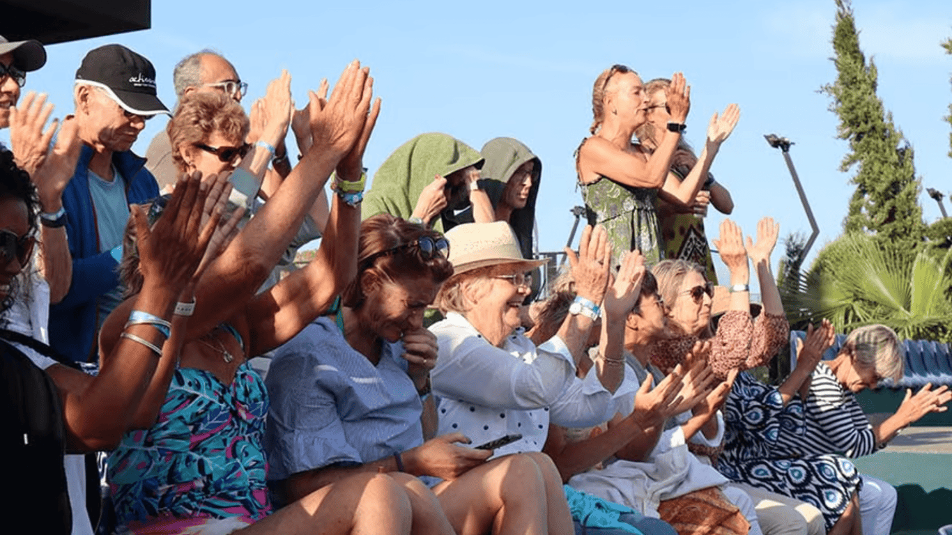 Crowd of holiday clients clapping and cheering courtside at a sunny tennis event.