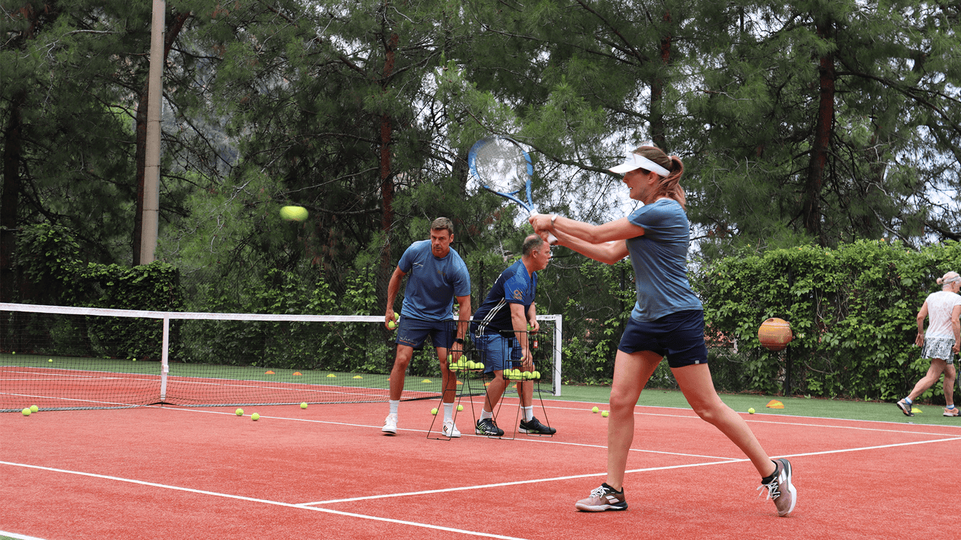 Woman hits a backhand during a cardio tennis drill as coaches feed balls on an outdoor court.