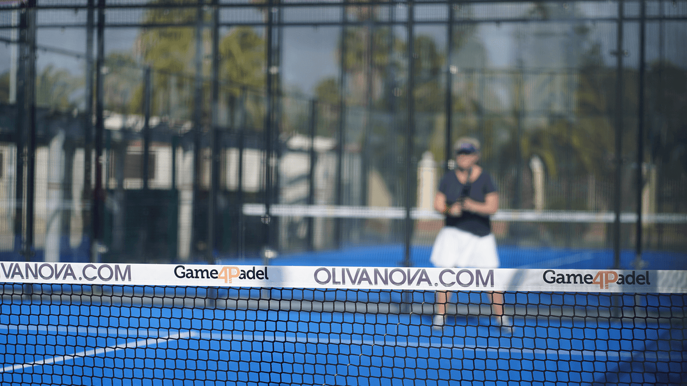 Padel net branded Game4Padel at Oliva Nova, with player blurred in background on blue court.
