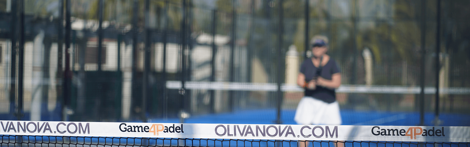 Padel net branded Game4Padel at Oliva Nova, with player blurred in background on blue court.