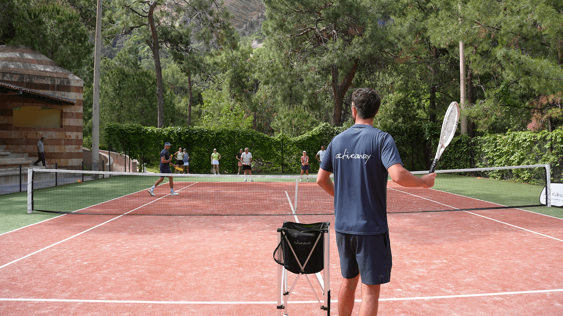 Tennis coach leads a lesson on an outdoor clay court as players practise and watch at Liberty Lykia.