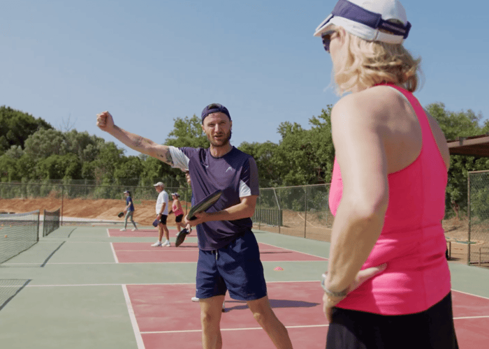Pickleball coach demonstrating a drill to a player on outdoor courts during a sunny coaching session.