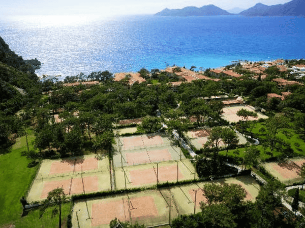Aerial view of seaside resort with multiple tennis courts among trees, overlooking a blue bay and mountains.