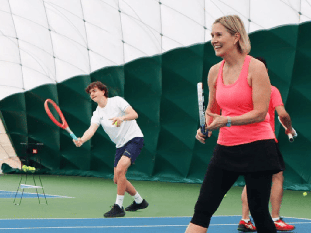 Smiling woman plays a tennis drill indoors as other players practise behind her.