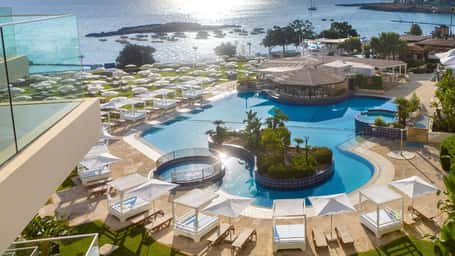 Aerial view of Capo Bay resort main pool with cabanas, sunbeds and the sea beyond at sunset.