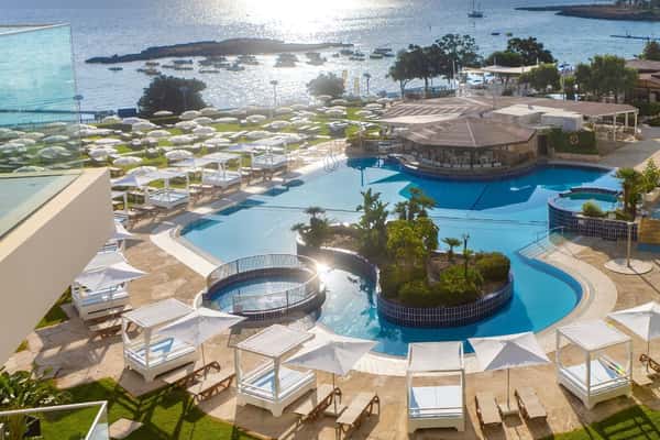 Aerial view of Capo Bay resort main pool with cabanas, sunbeds and the sea beyond at sunset.