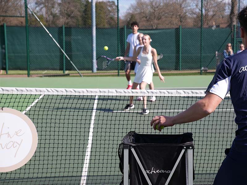 Coach feeds balls to two junior players during an outdoor tennis drill on a sunny court.
