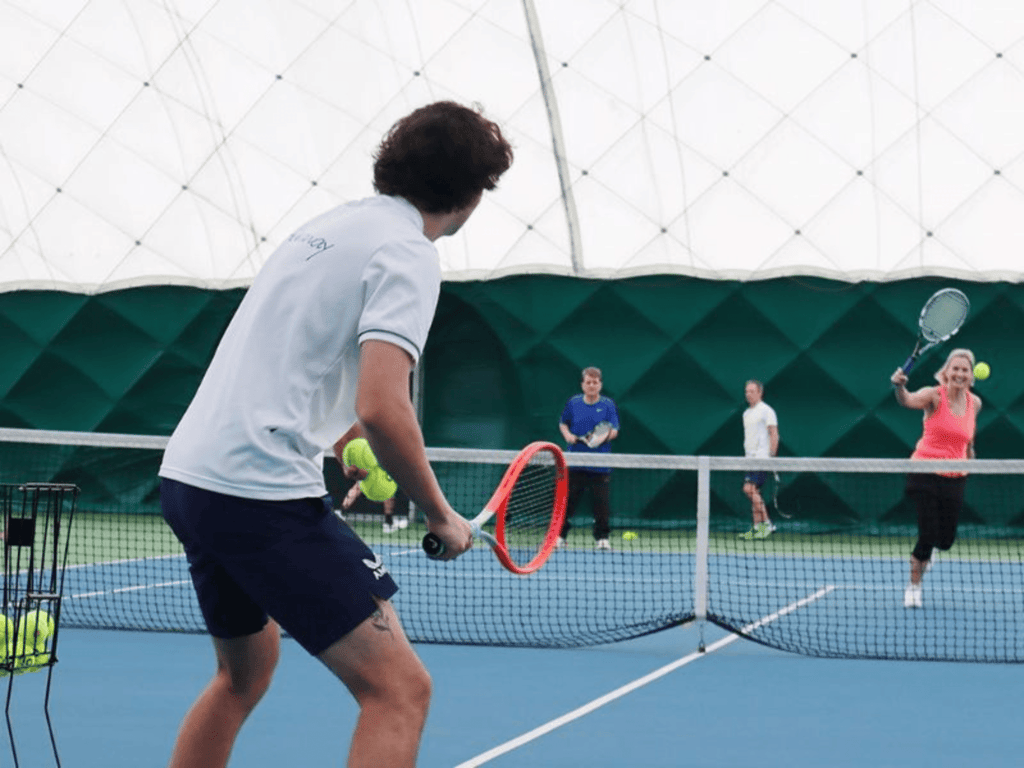 Coach and players practise at an indoor tennis clinic, rallying across the net.