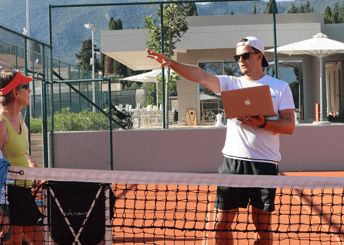 Tennis coach with laptop instructs two players by the net on a sunny outdoor court.